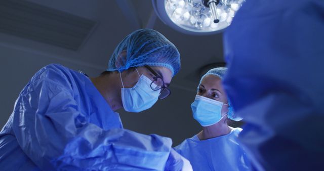 This image showcases a team of surgeons dressed in blue scrubs and wearing masks, performing an operation under bright surgical lights in an operating room. It portrays a sterile environment typical of hospitals, emphasizing the professionalism and meticulous nature of healthcare workers. Ideal for use in healthcare promotions, educational materials, articles on medical procedures, or advertisements for medical services.