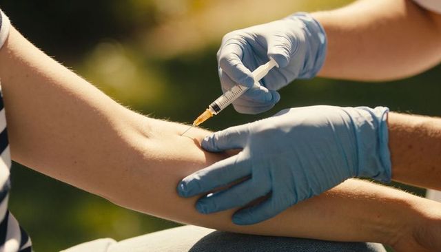 Healthcare worker injecting vaccine into forearm outdoors wearing blue gloves striped shirt