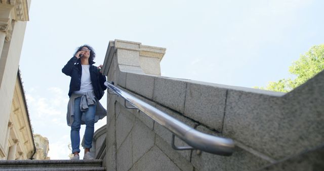 Casual woman walking up stone staircase while talking on smartphone. Denim jacket tied around waist, casual attire. Possible uses include articles on communication, urban lifestyle, outdoor activities, and casual fashion.