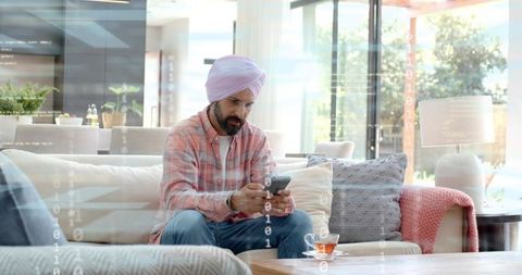 Indian man wearing pink turban tapping smartphone in modern lounge with digital overlay