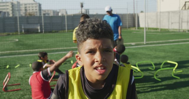 A young football player with dark curly hair wearing a yellow training bib resting during an outdoor training session. Others are interacting and aiding in the background. Ideal for content related to youth sports, teamwork, fitness, and athletic training.
