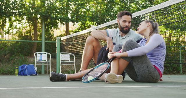 Couple sitting on tennis court resting. Man and woman leaning against net, holding tennis racket and ball. Summer day with sunlight filtering through trees in background. Ideal for promoting sportsmanship, fitness activities, outdoor recreation, and leisure marketing.