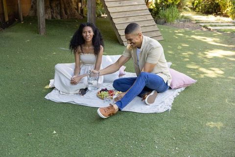 Relaxed couple sharing backyard picnic on blanket pouring water with fresh fruit platter