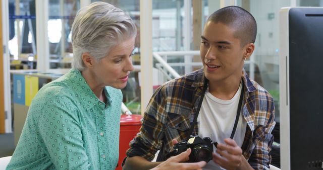 Caucasian woman and young biracial man review a camera in an office. They're collaborating on a project, sharing insights and expertise.