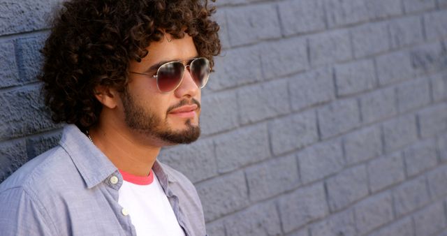 Pensive biracial man with curly hair and sunglasses, looking ahead by wall on sunny street. City break, summer, travel, vacations and lifestyle, unaltered.