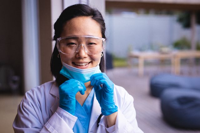 This image depicts a smiling Asian doctor wearing protective glasses and a face mask, symbolizing safety and professionalism in healthcare. Ideal for use in medical articles, health service promotions, COVID-19 awareness campaigns, and educational materials about healthcare workers and their roles during the pandemic.