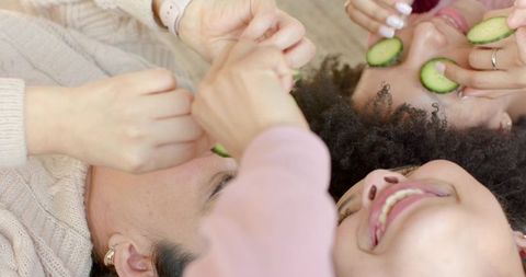 Multiracial friends laughing while holding cucumber slices during cozy home spa moment