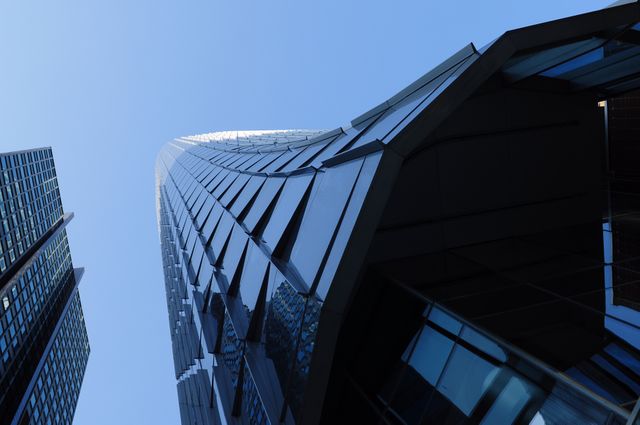 Photo depicts upward view of a modern skyscraper with reflective glass facade against clear blue sky. Suitable for use in urban-themed presentations, architectural design websites, or corporate branding materials highlighting modernity and advancements in construction.