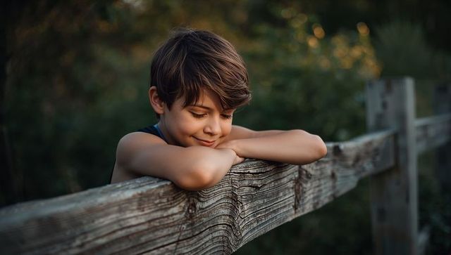 Boy leaning on weathered wooden fence rail reflecting at golden hour in countryside