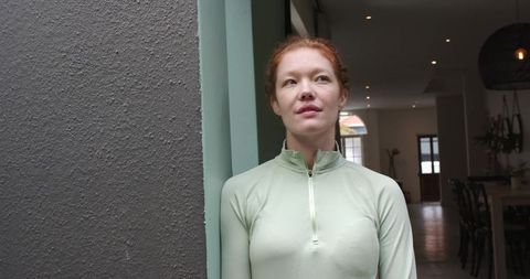 Contemplative red-haired woman standing in doorway wearing seafoam zip-up top minimalist open-plan