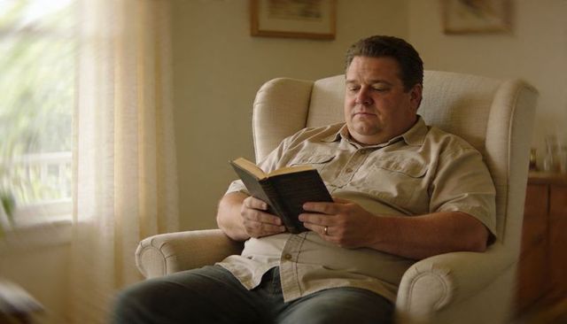 Middle-aged man reading paperback in sunlit armchair cozy home reading nook