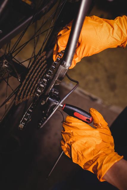 Mechanic wearing orange gloves repairing a bicycle chain in a workshop, focusing on precision and skill. This image can be used for content related to bike maintenance, mechanical skills, cycling, and professional repair services.