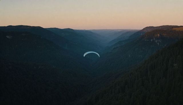 Gliding paraglider drifting over dusk forested mountain valley with pale canopy and pilot