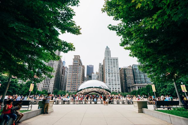 Tourists gather around the iconic Cloud Gate sculpture in Chicago's Millennium Park, with a backdrop of tall city buildings. Ideal for illustrating urban tourism, travel destinations, city landmarks, or public spaces.