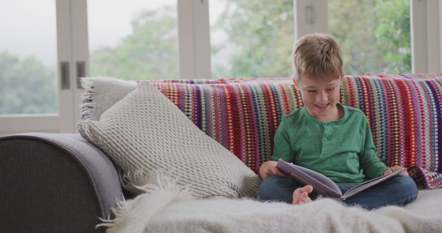 A young boy in casual clothes is sitting on a comfortable sofa covered with a colorful blanket, attentively reading a storybook. Natural light filters through large windows in the background, creating a warm and inviting environment. Perfect for use in promotions related to childhood education, reading habits, home comfort, and children's activities.