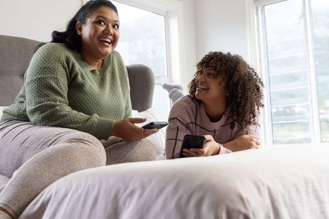 Hispanic mother and african american teenage daughter laughing and bonding on bed while using smartp