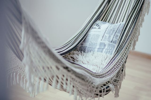 Hammock with fringe detail suspended indoors. A pillow with newspaper print and a soft, furry blanket add to the comfortable and stylish setting. Ideal for home decor ideas, relaxation concepts, articles on creating cozy spaces, or inspiration for indoor leisure activities.
