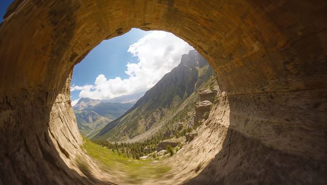 Circular rock tunnel framing sunlit alpine valley with arched opening and mountain peaks