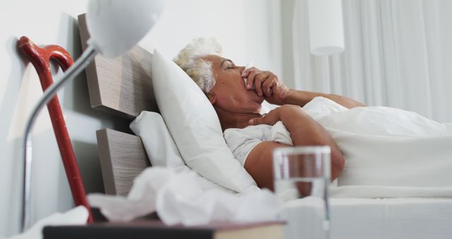 Elderly woman lying in bed with a cough, a walking cane by her bedside, and a glass of water nearby. Photo can be used to represent healthcare, illness, senior care, medical treatment, and home care scenarios involving elderly individuals.