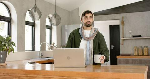 Indian man working remotely at kitchen island with laptop and coffee in modern rustic home