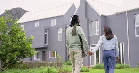African american and indian couple walking hand-in-hand toward modern gray house on lawn