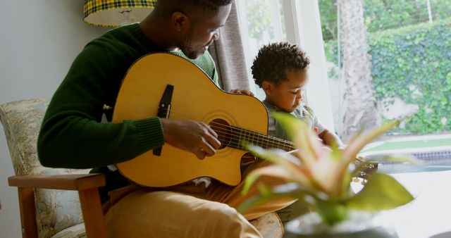 Father bonding with young son while teaching him to play acoustic guitar in cozy living room. Themes of parent-child relationship and music education. Perfect for use in parenting blogs, educational content, music-related advertisements, family-oriented promotions, and home interior articles.