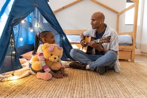 African american father playing guitar while daughter hugging teddy bear in cozy teepee bedroom