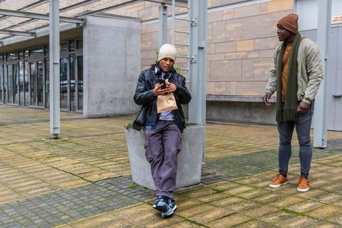 Two diverse men leaning on planter in urban plaza scrolling smartphone and chatting
