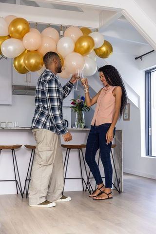 Young couple toasting with wine in sunlit modern kitchen under gold and blush balloons