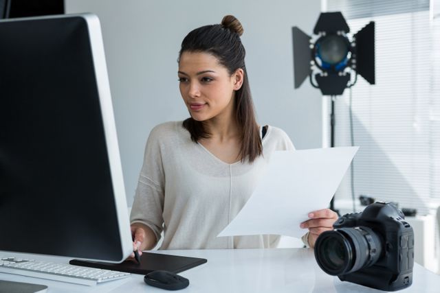 Young female photographer working in a modern studio, using a graphic tablet and computer for photo editing. Ideal for use in articles or advertisements related to photography, digital editing, creative professions, and modern workspaces.