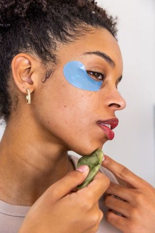African american woman using gua sha and blue under-eye patch for glowing skincare routine