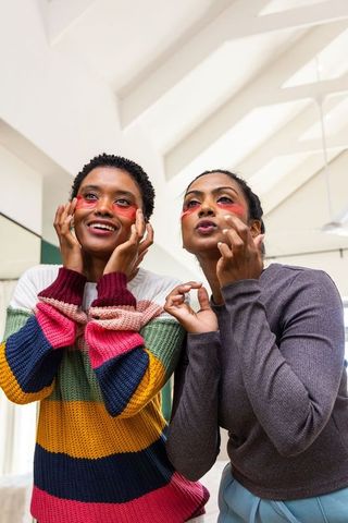Indian and african american friends applying red under-eye patches smiling while grooming