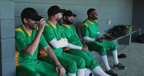 Baseball teammates laughing and preparing in dugout wearing green and yellow uniforms