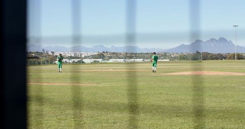 Two cricketers in green uniforms standing on outfield behind fence with mountain backdrop