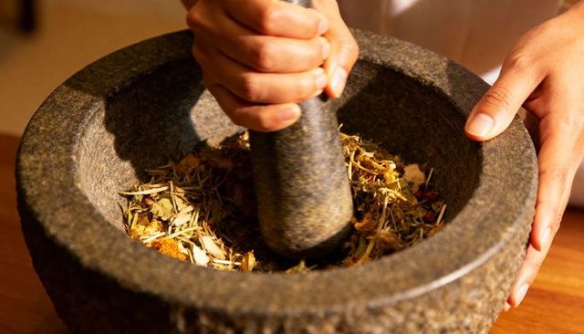 Hands Grinding Dried Herbs in Stone Mortar with Pestle on Wooden Counter