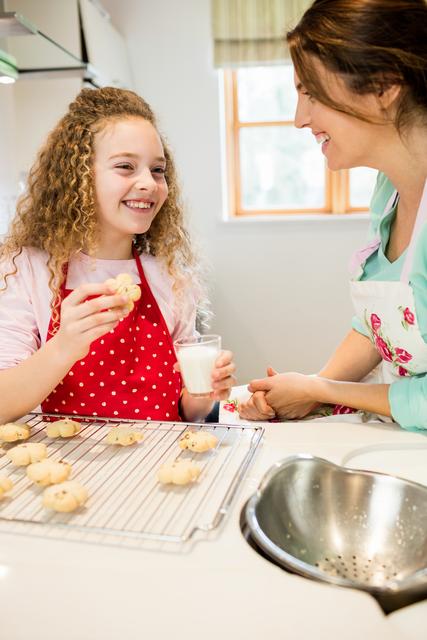 Mother and daughter enjoying quality time baking cookies in the kitchen. The daughter is holding a cookie and a glass of milk, while both are smiling and interacting. This image can be used for family-oriented content, cooking blogs, parenting articles, or advertisements promoting family activities and home baking.