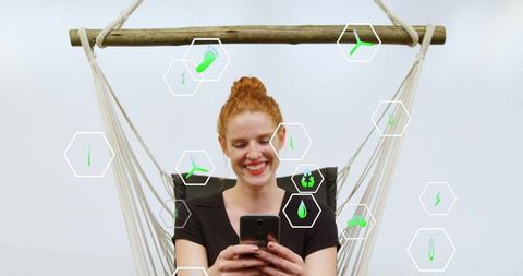 Smiling redhead woman in hammock using smartphone with green eco hex icons overlay