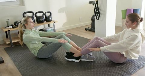 Two women performing partner core workout on mat in bright home gym with kettlebells