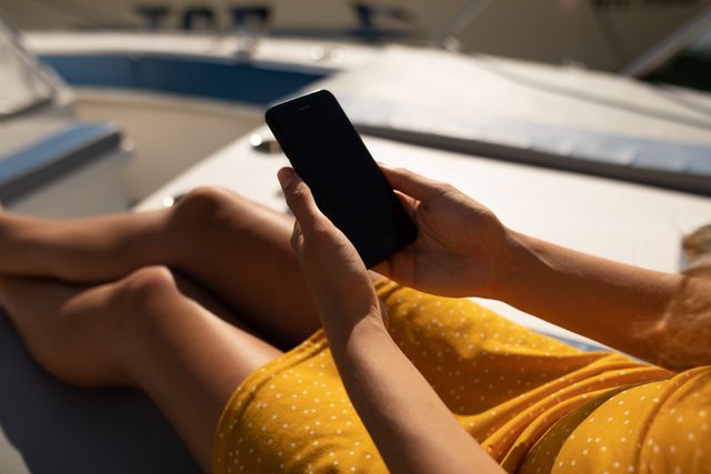 Teenage girl in a yellow dress is sitting on a boat by the sea on a sunny day, using her smartphone. This image can be used for themes related to summer vacations, leisure activities, technology use, and outdoor lifestyles. It is ideal for travel blogs, lifestyle magazines, and advertisements promoting summer fashion or mobile technology.
