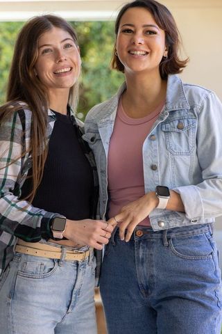 Two friends smiling while showing smartwatches and rings on porch in casual denim
