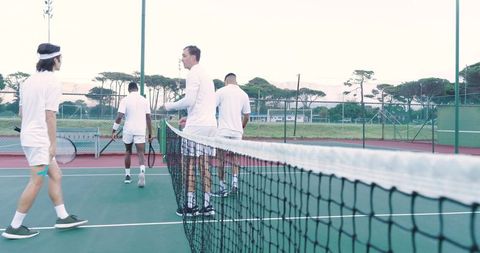 Four male tennis players walking past net wearing white outfits on outdoor hardcourt