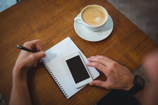 Man writing notes on notepad while using smartphone in cafe. Coffee cup on table. Ideal for illustrating concepts of productivity, business planning, casual work environment, and modern technology use.