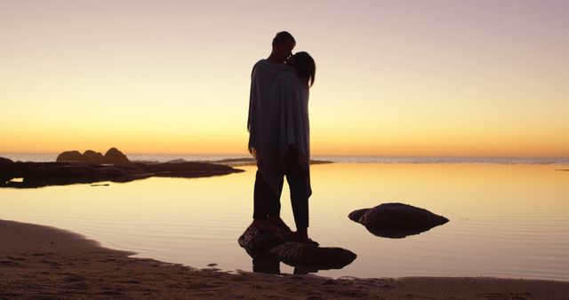 Silhouetted couple standing and embracing on a rock in shallow water during sunset at the beach. Used for themes related to romance, intimacy, nature, serenity, and tranquility. Perfect for greeting cards, romantic scenes, lifestyle blogs, travel brochures.