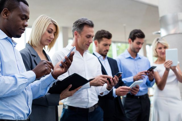 Group of business executives standing in a line, using digital tablets and mobile phones at a conference center. Ideal for illustrating concepts of modern business communication, technology in the workplace, corporate meetings, and professional networking.