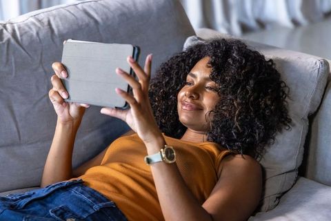 Woman reclining on sofa relaxing while using tablet for streaming and reading
