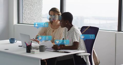 Two women reviewing documents at office desk with laptop and social media notifications
