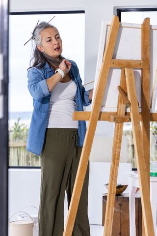 Senior woman painting at easel in bright studio wearing denim shirt and relaxed green pants