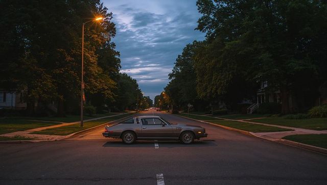 Vintage coupe sitting across dusk suburban street under glowing lampposts and tree canopy