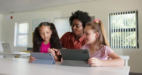 Diverse female teacher guiding two girls using tablets in bright classroom collaboration