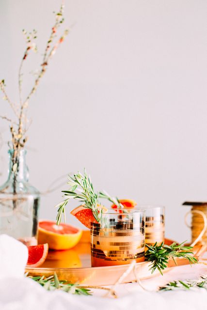 Winter-themed cocktail featuring grapefruit and rosemary in stylish gold rim glasses. The grapefruit and rosemary provide a fresh, festive touch amidst other stylish table elements. Perfect for food and drink blogs, holiday drink ideas, and social media posts about elegant beverages.